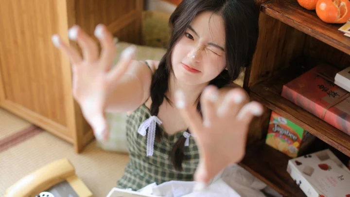 一名身穿绿色格纹吊带裙的女性在木质室内对着镜头眨眼并伸出双手。