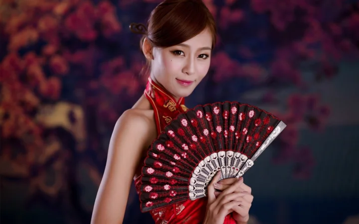 一名成年女子身穿红色服装，手持装饰华丽的折扇面对镜头。