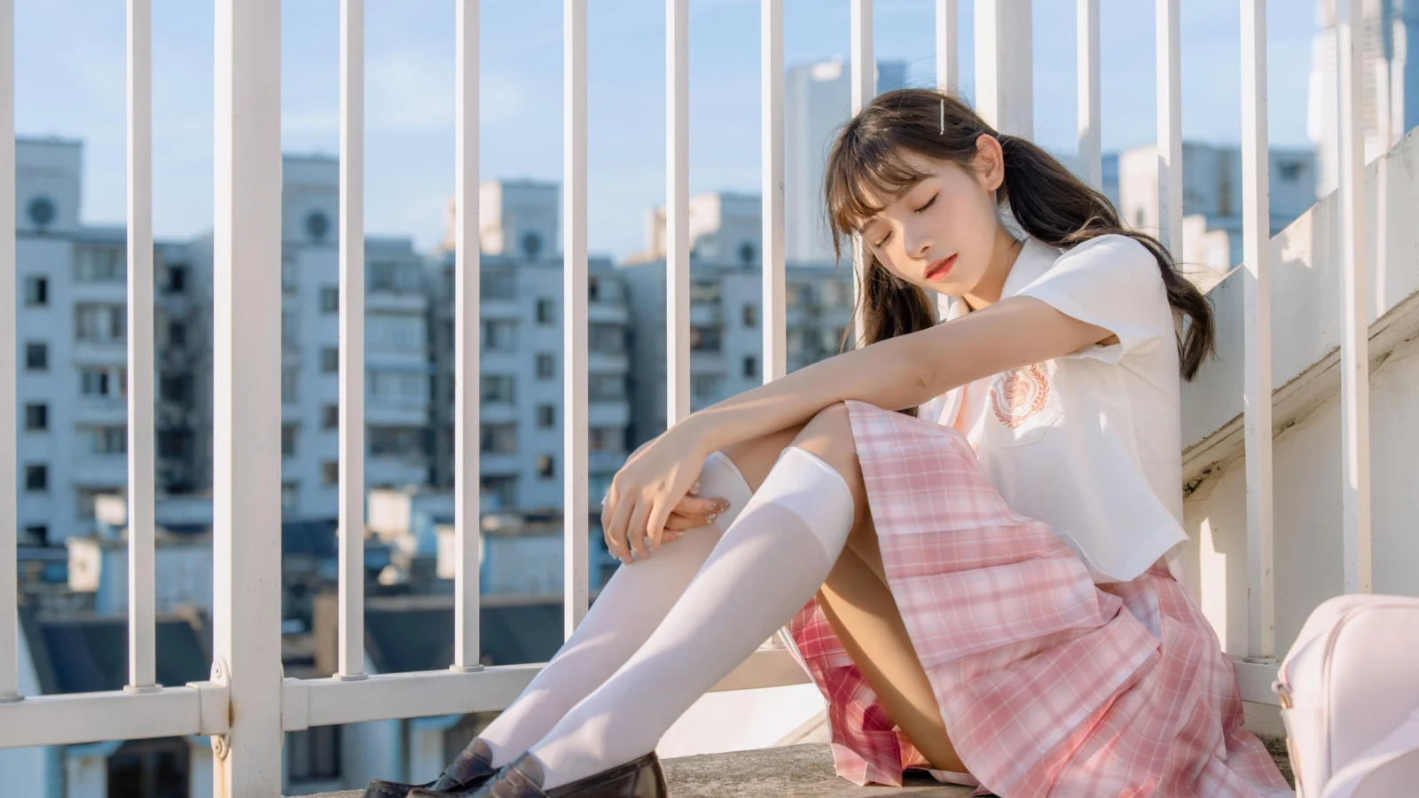 젊은 여성이 발코니 난간 옆에 앉아 있고 뒤로 도시 건물이 보입니다.