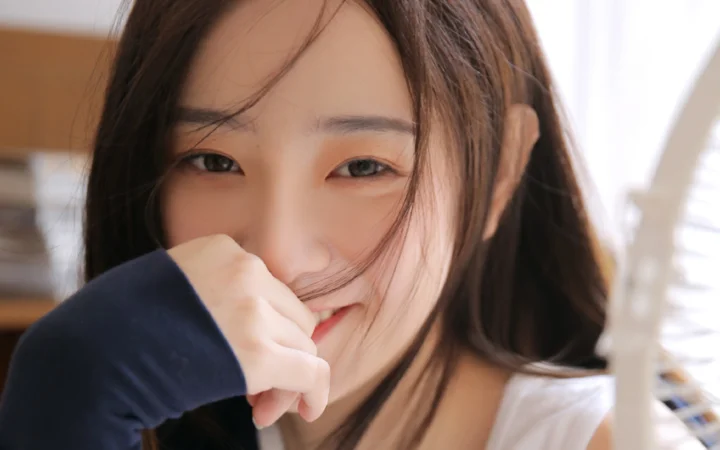 一名长发女性在室内靠近镜头微笑，手臂遮在嘴边。