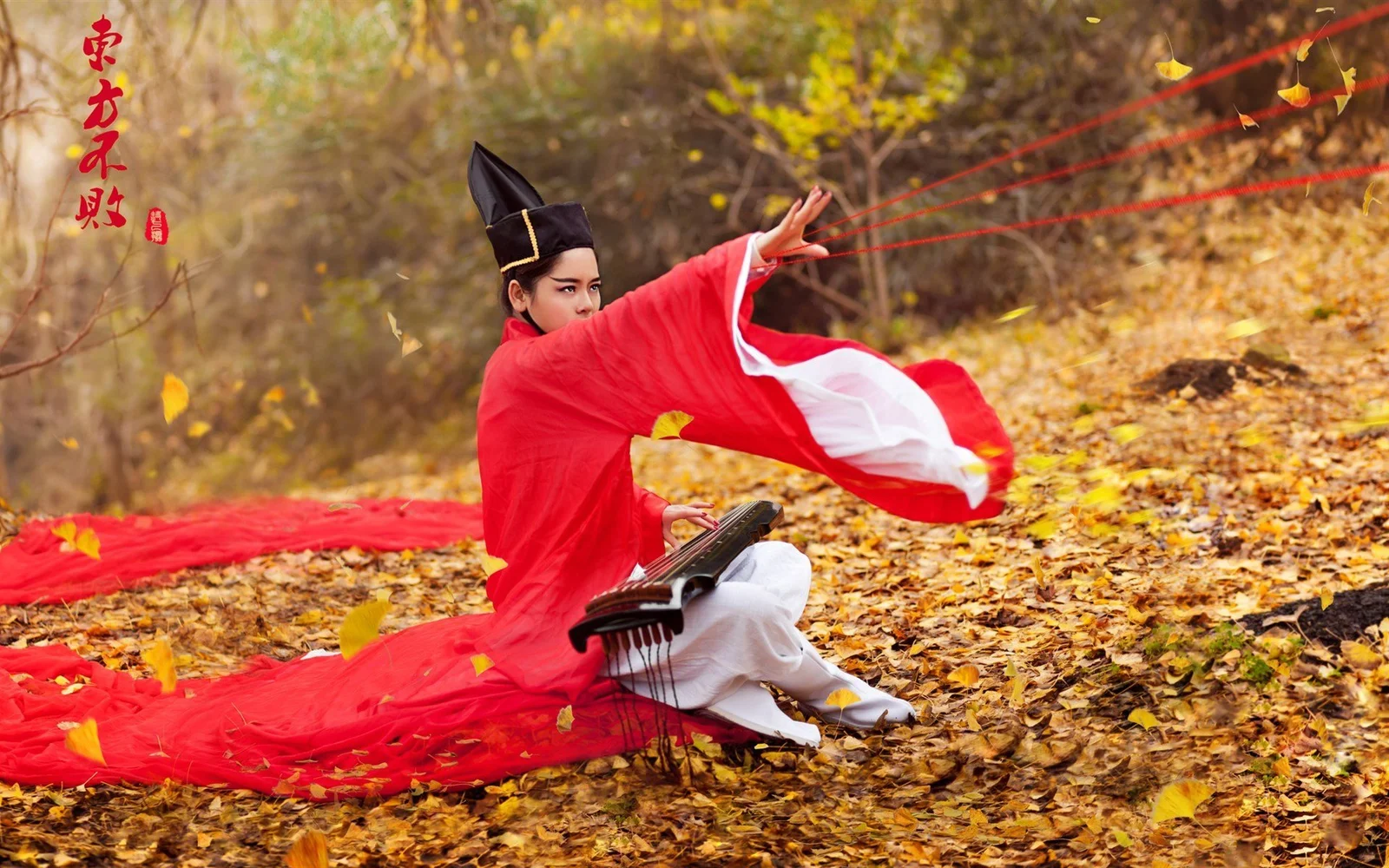 一名身穿红色传统风格服饰的女子坐在秋叶间，身旁放着古琴般的弦乐器，红色衣料随风展开。