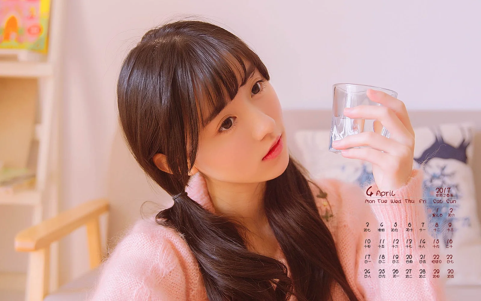 분홍색 상의를 입은 성인 여성이 실내에서 유리잔을 들고 있으며 배경에 달력이 보입니다.