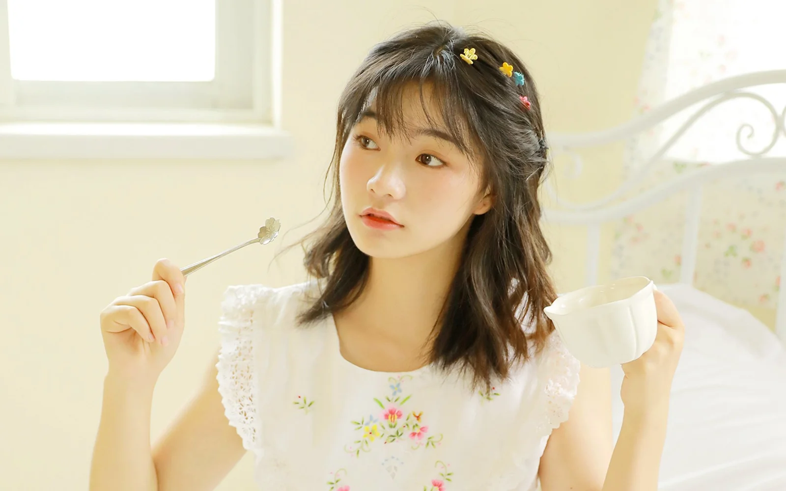 花のヘアアクセサリーを付けた女性が、明るい室内でティーカップと小さなスプーンを持っています。