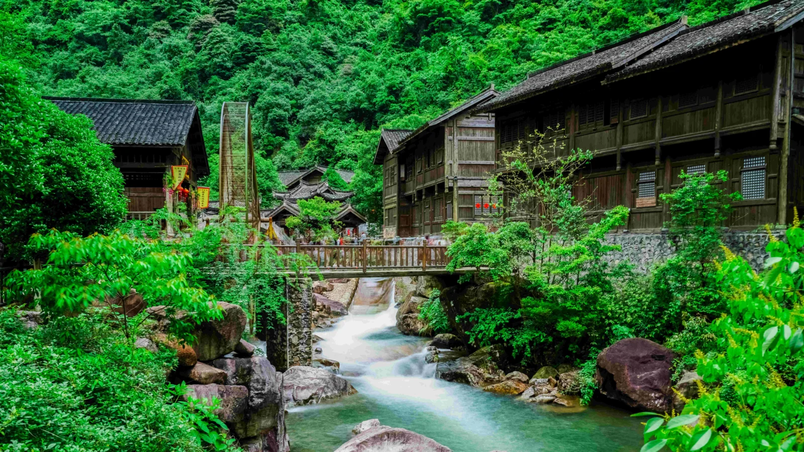 茂密森林中的木屋村落旁，一条清澈溪流从桥下流过。