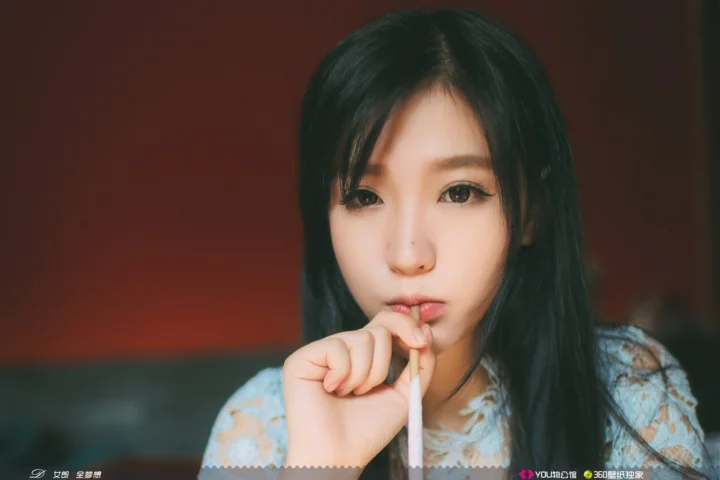 一名黑发女性面对镜头，手指轻触嘴唇，背景为深色室内环境。