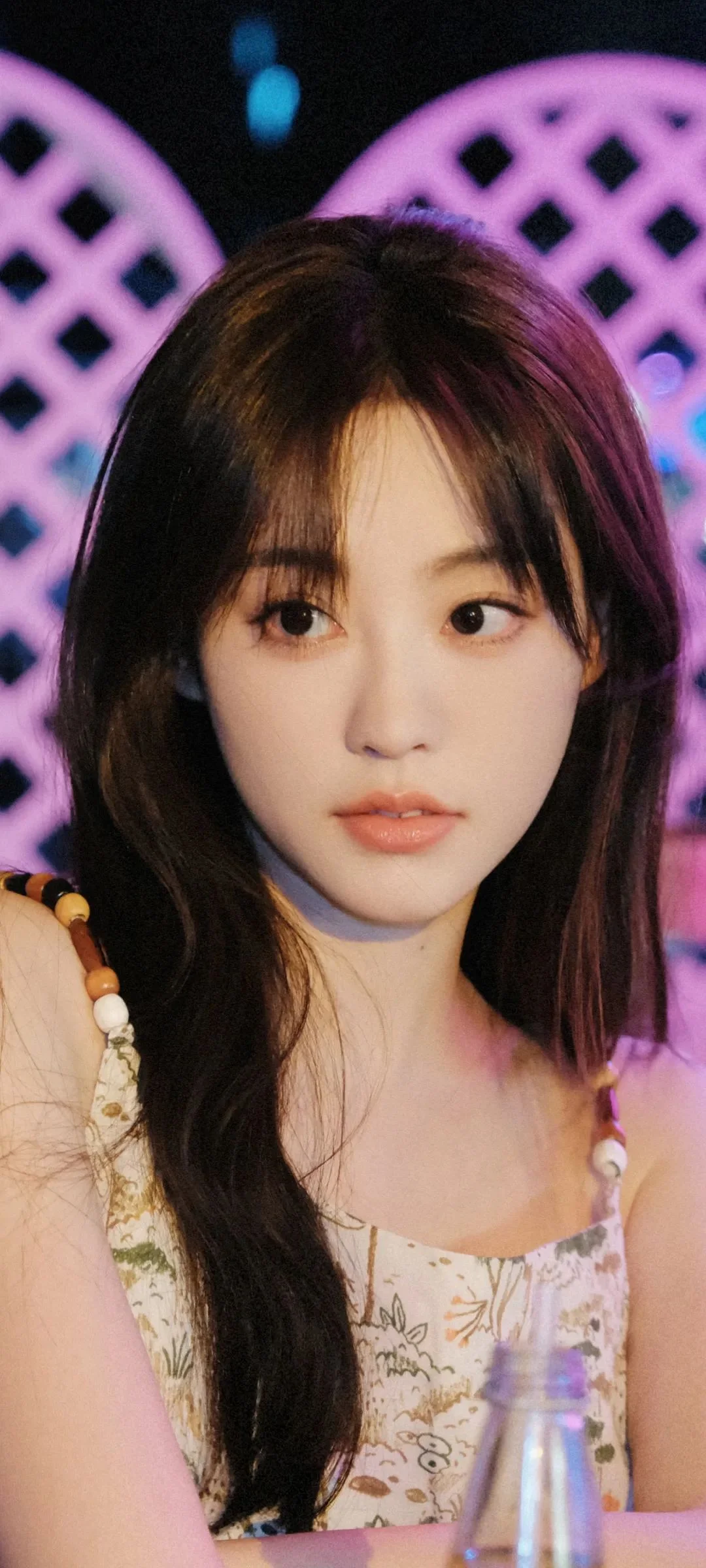 긴 머리의 여성이 꽃무늬 상의를 입고 보라색과 파란색 조명 무늬를 배경으로 카메라를 바라보고 있습니다.