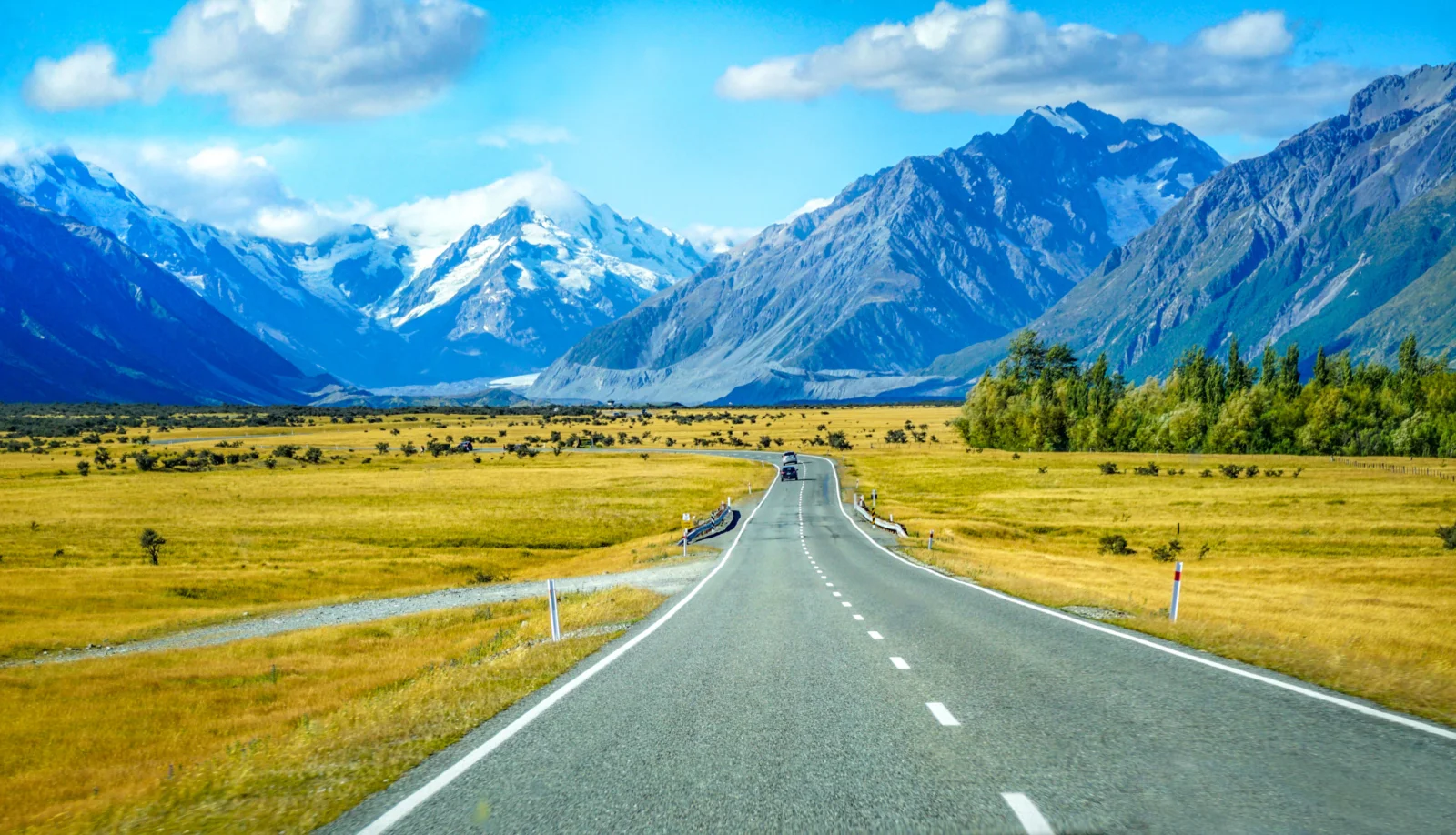 一条公路穿过金色草原，通向被蓝天白云环绕的壮丽雪山。