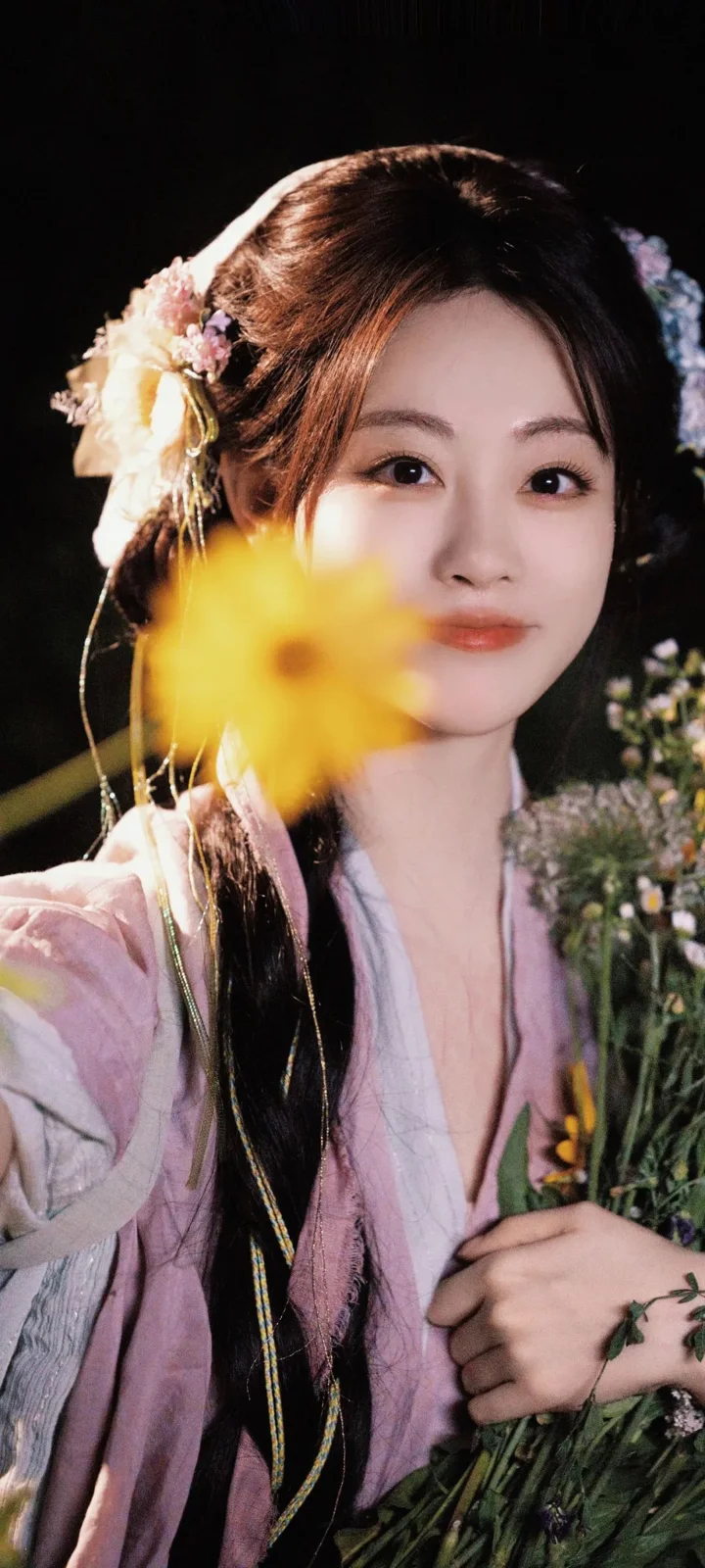 一位身穿淺粉色傳統服飾的女子手持花束，面前有一朵黃色花朵虛化入鏡。