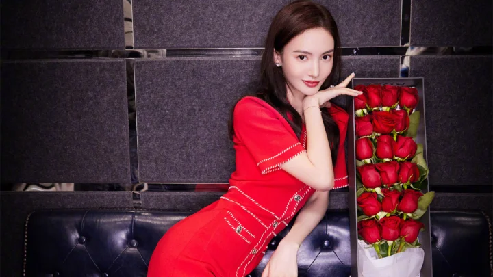 一名身穿红色连衣裙的成年女子坐在深色沙发旁，身边放着一束红玫瑰。