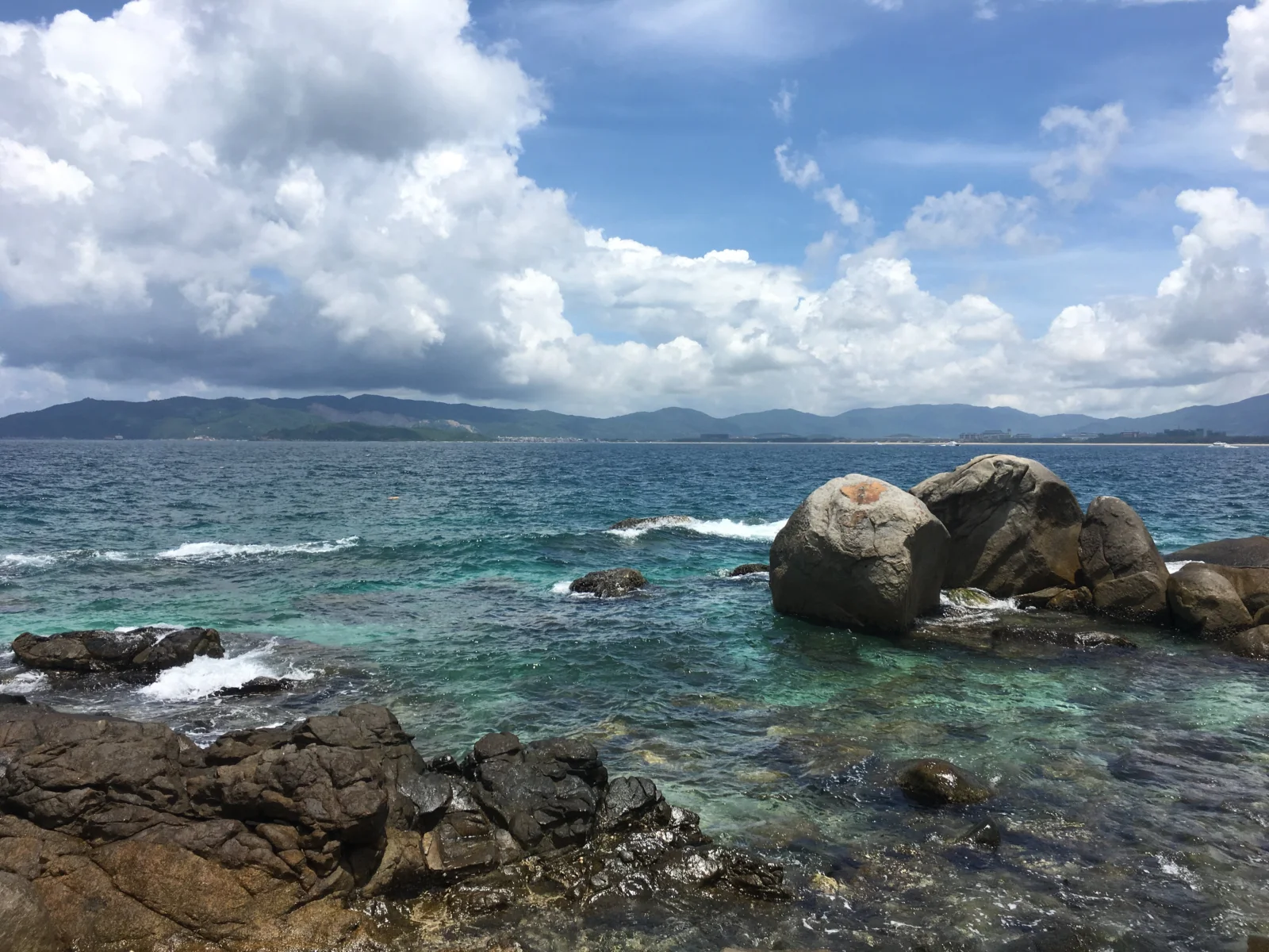 清澈的海水拍打着礁石，远处是连绵山峦，天空布满蓬松的白云。