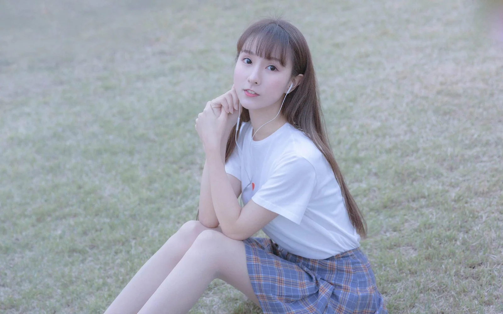 一名成年女性坐在草地上看向鏡頭，穿著白色上衣和格紋裙。