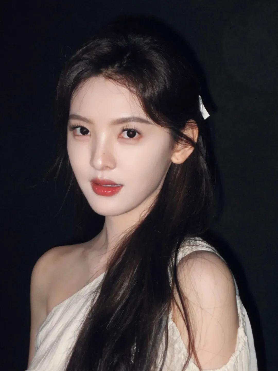 검은 배경 앞에서 흰색 상의를 입은 긴 머리 여성이 카메라를 바라보고 있습니다.