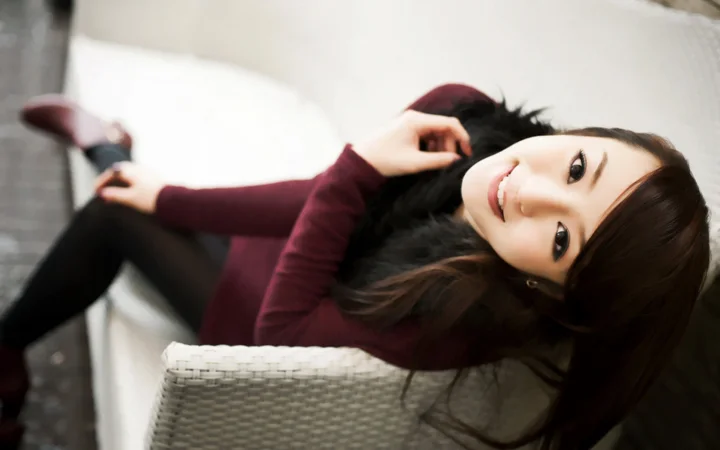一名棕发女性穿着酒红色上衣和黑色裤袜，斜躺在编织椅上微笑看向镜头。
