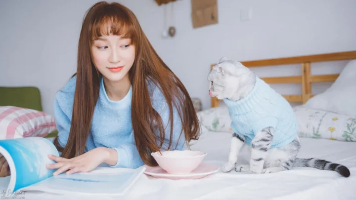 一名长发女子坐在床上，旁边是一只穿着浅蓝色衣服的猫咪。