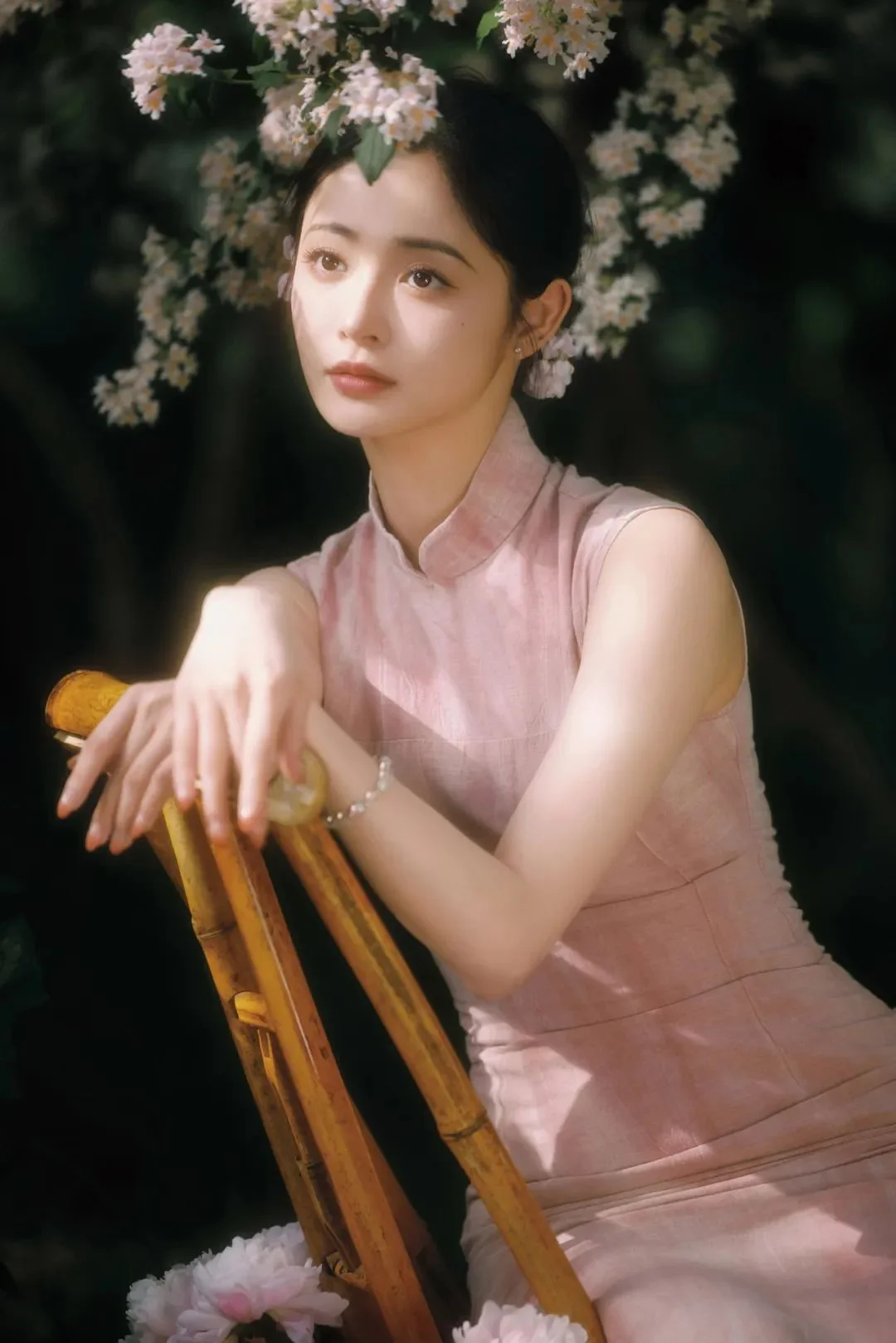 연한 꽃들에 둘러싸인 어두운 배경에서 분홍색 민소매 드레스를 입은 여성이 대나무 의자 옆에 앉아 있습니다.