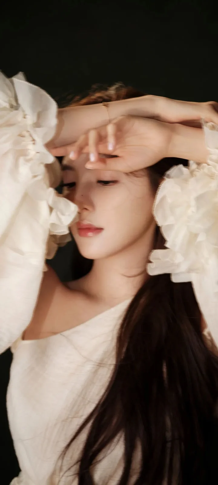 긴 머리의 여성이 흰색 드레스를 입고 어두운 배경 앞에서 팔을 이마에 올리고 있다.