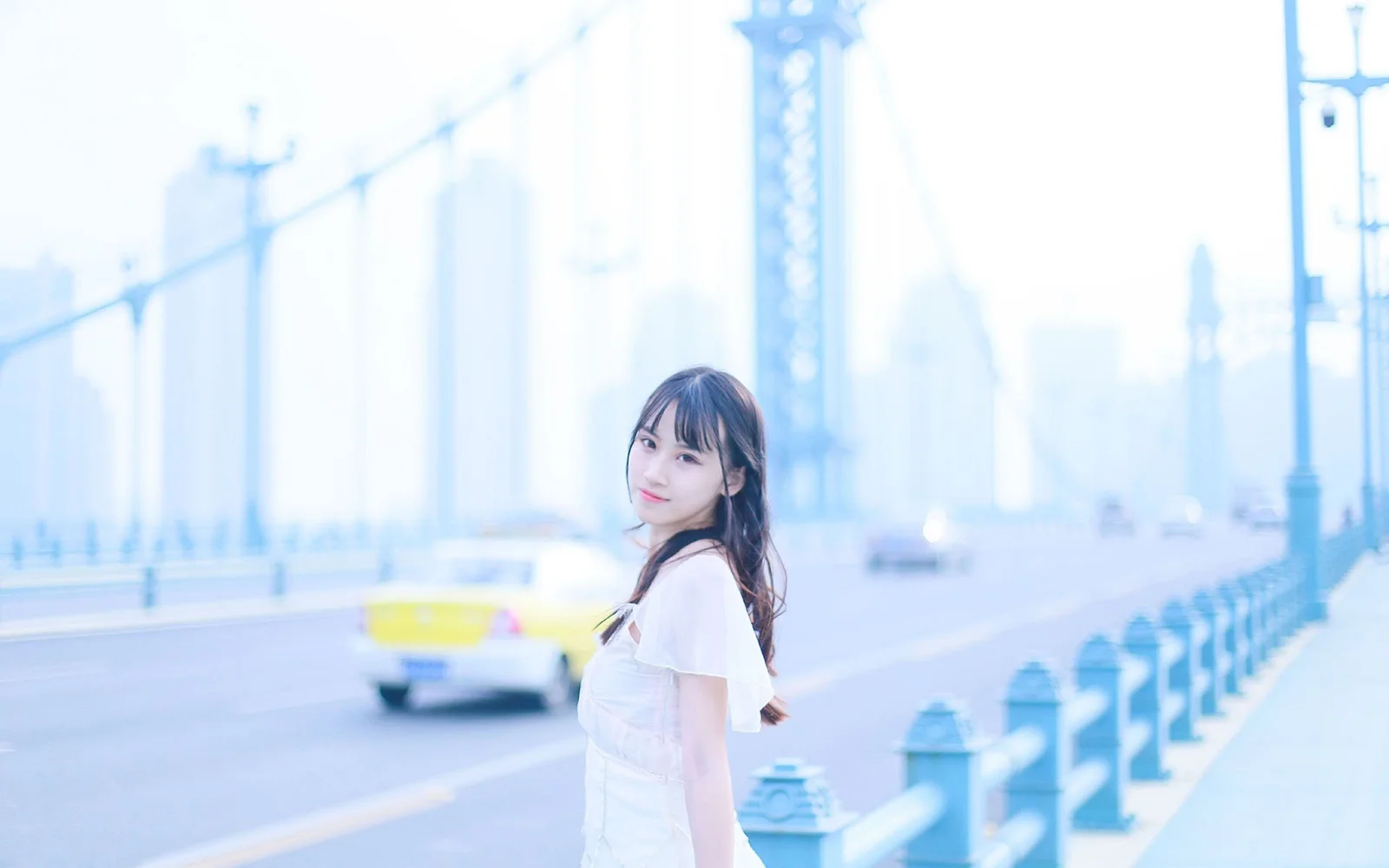 女性が都市の橋の歩道でカメラを振り返り、背後に明るい道路や欄干、高層ビルの輪郭が見える。