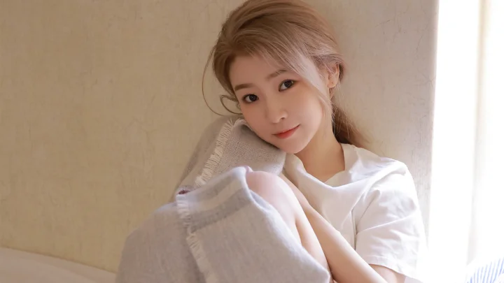 성인 여성이 실내 창가에 앉아 밝은 색 숄을 두르고 부드러운 빛을 받고 있습니다.