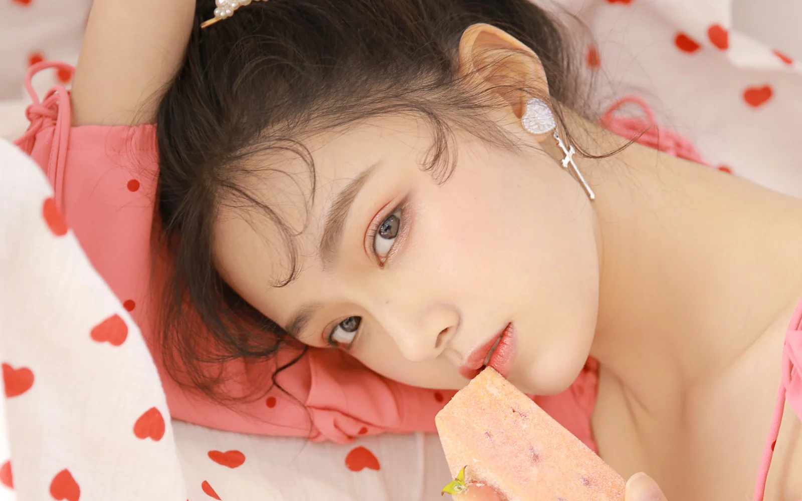 빨간 하트 무늬의 분홍색 침구 위에서 여성이 분홍색 아이스바를 입가에 대고 있습니다.