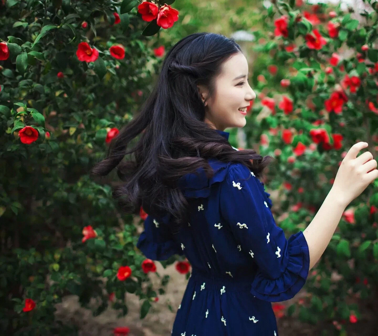 짙은 파란색 원피스를 입은 여성이 붉은 꽃이 핀 정원에서 돌아보며 미소 짓고 있다.