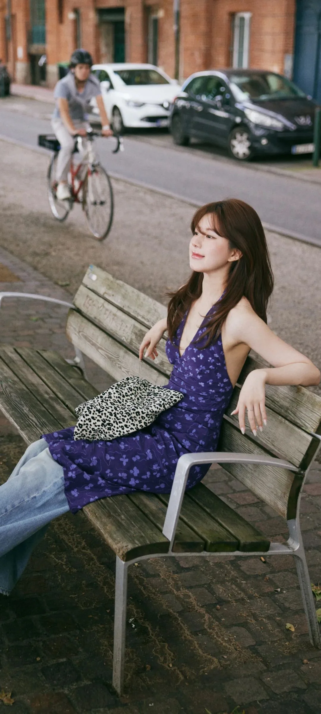 한 여성이 도시 거리 옆 벤치에 앉아 있고 뒤로 자전거와 자동차, 건물이 보인다.