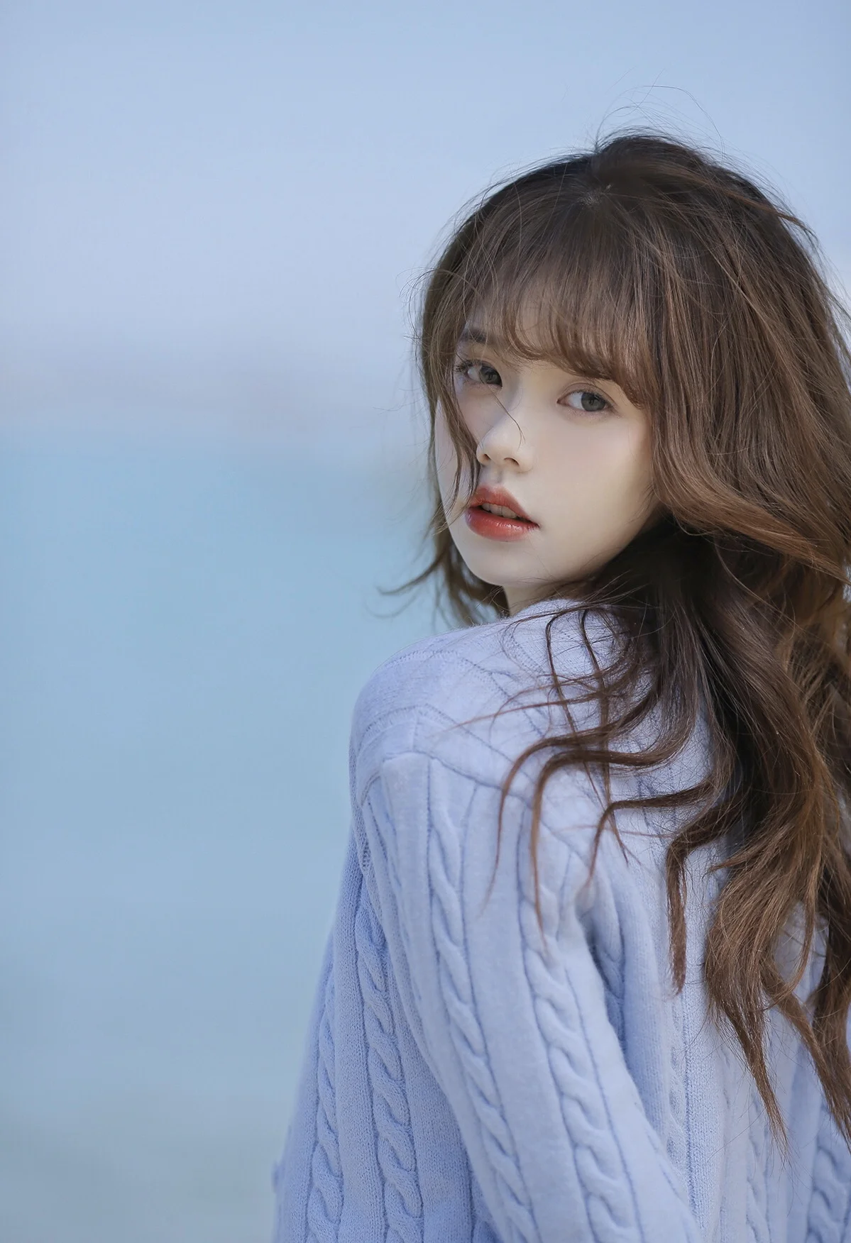 긴 머리의 여성이 연한 파란색 니트를 입고 파란 배경 앞에서 뒤돌아 카메라를 바라보고 있다.