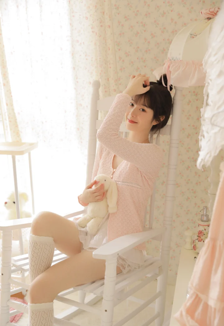 ピンクのニット服を着た女性が白い椅子に座り、淡い色のぬいぐるみを抱えています。