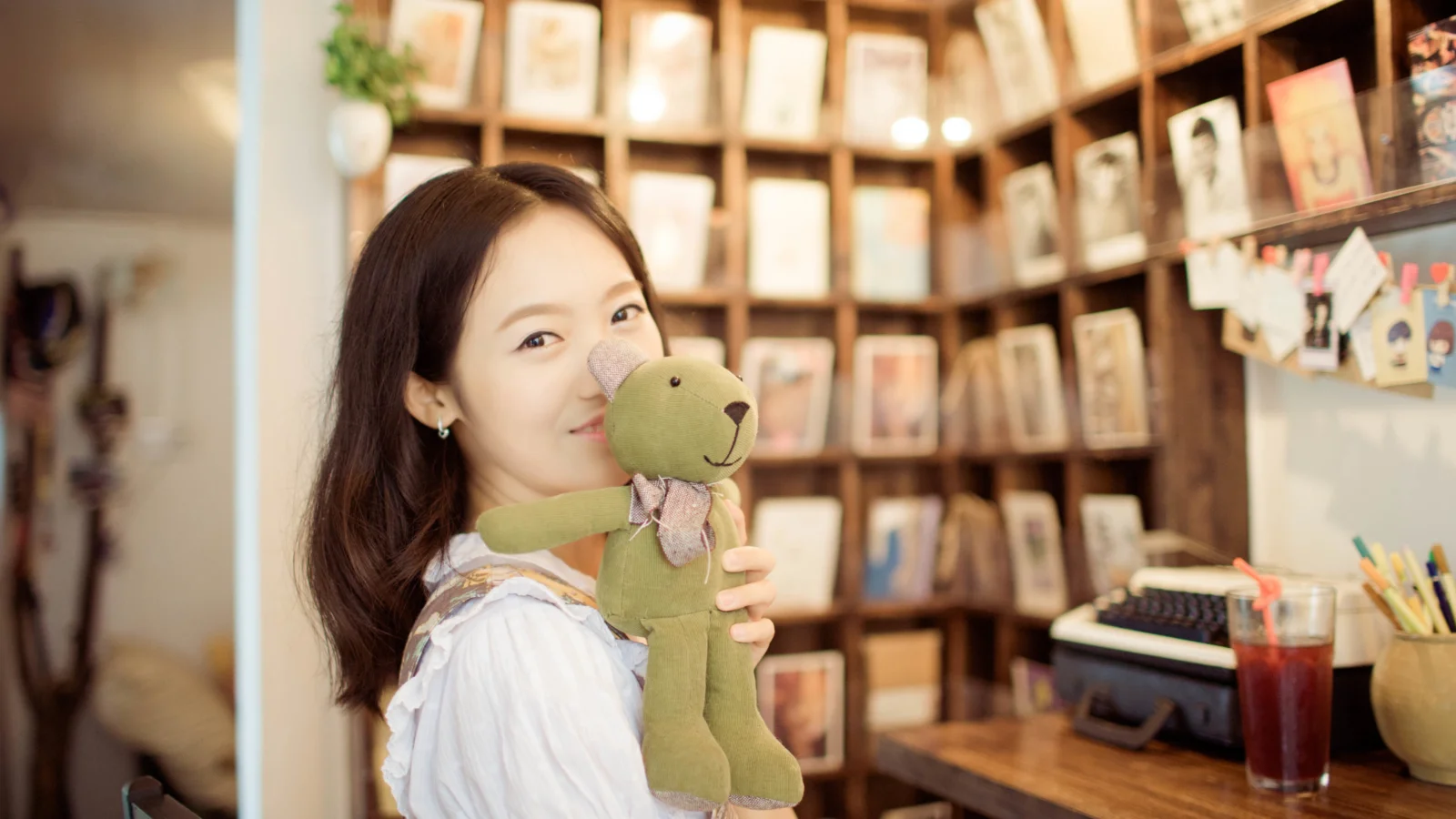 실내에서 성인 여성이 초록색 봉제 인형을 안고 있으며 뒤에는 액자와 물건이 놓인 나무 선반이 있습니다.