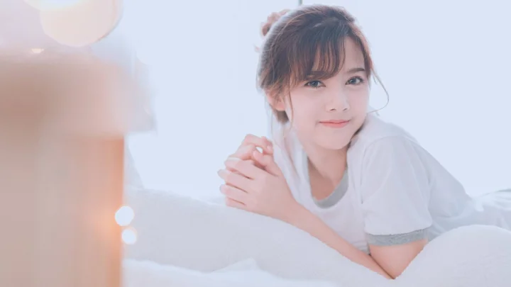 白いシャツを着た女性が白い寝具の上でカメラに向かって微笑んでいます。