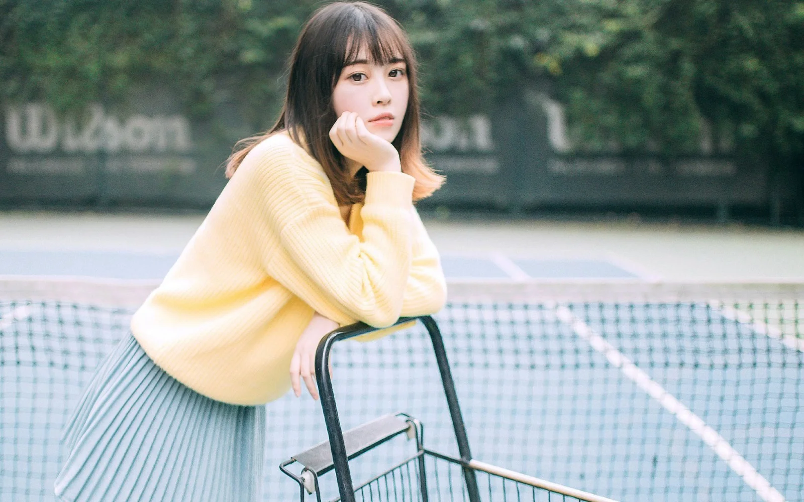 一名穿著淡黃色毛衣的女子靠在網球場邊的金屬推車旁。