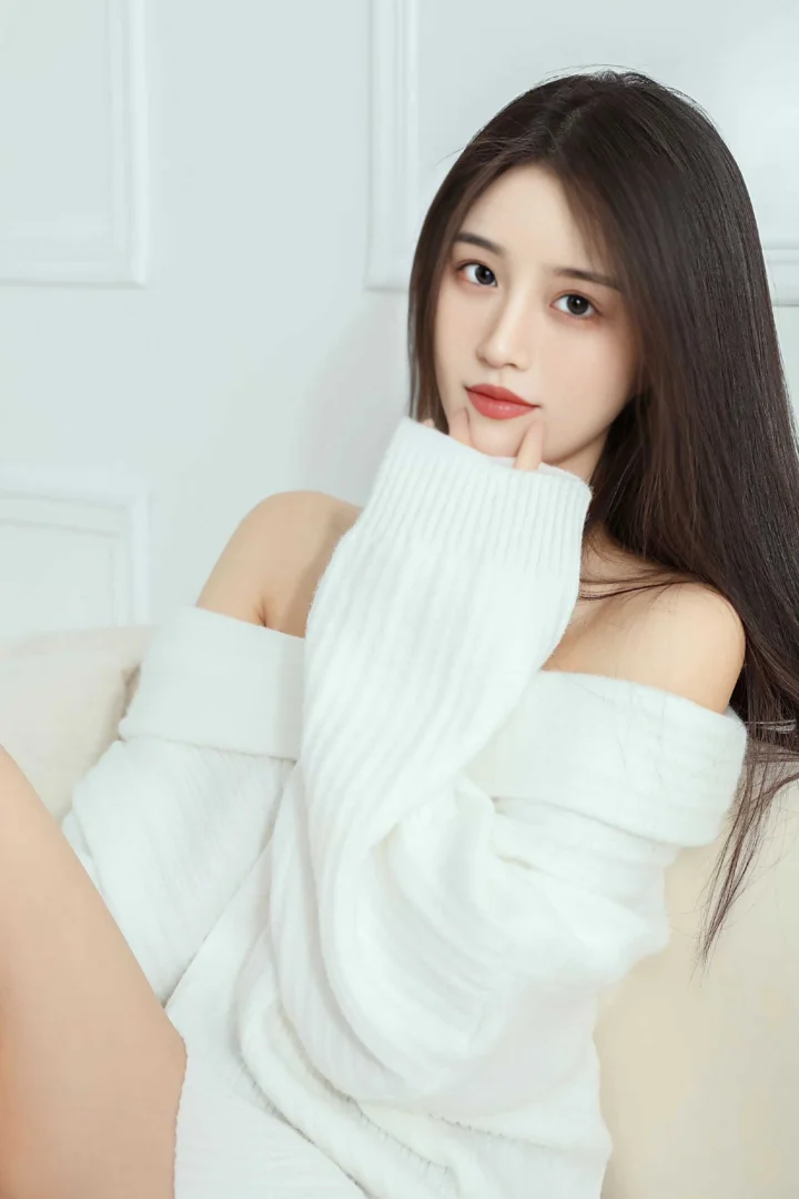 긴 머리의 여성이 흰색 니트를 입고 실내에 앉아 카메라를 바라보며 부드럽게 미소 짓고 있다.