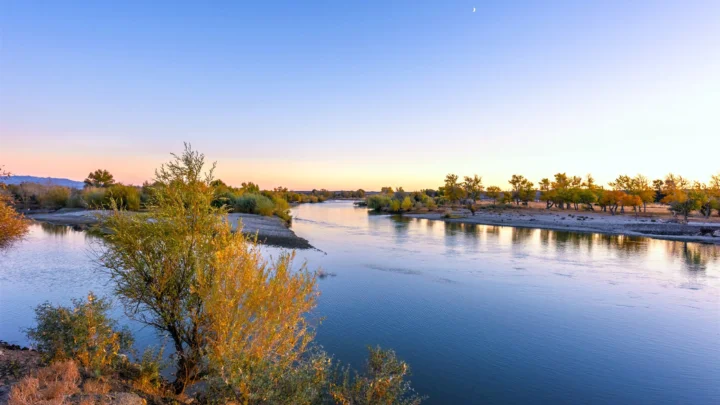 夕阳映照下的河流穿过两岸树木与沙洲，天空呈现柔和的蓝紫与金色渐变。