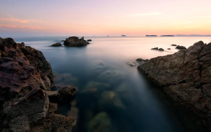 暮色天空下，海水环绕着前景中的深色礁石海岸，远处可见几座小岛。