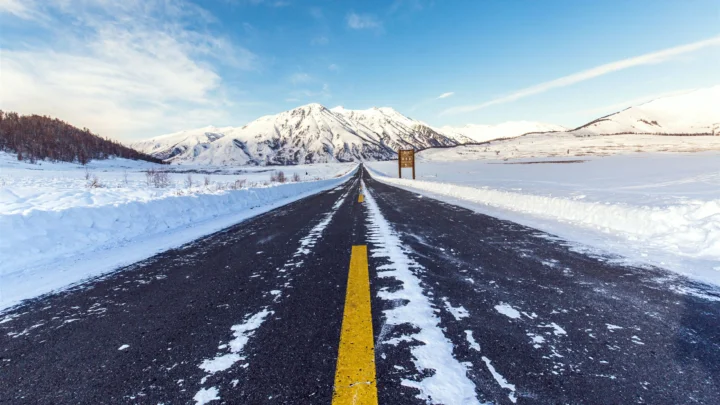 一条覆有积雪的公路穿过白色雪原，远处是壮丽的雪山和晴朗的蓝天。
