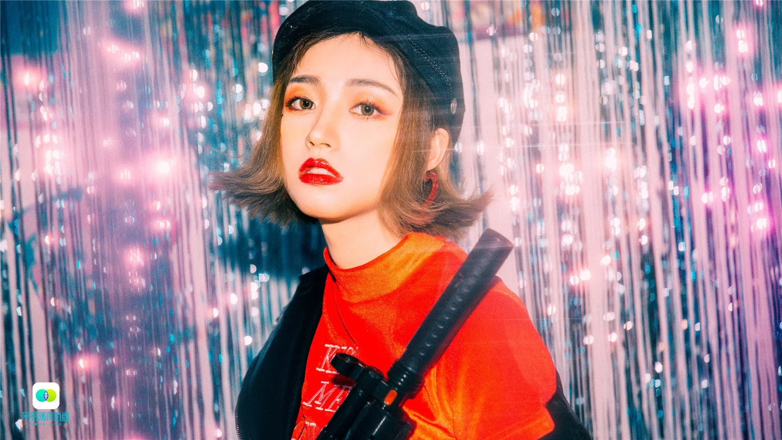 검은색 헤드웨어와 빨간 상의를 착용한 성인 여성이 분홍색과 파란색으로 반짝이는 커튼 앞에 서 있습니다.