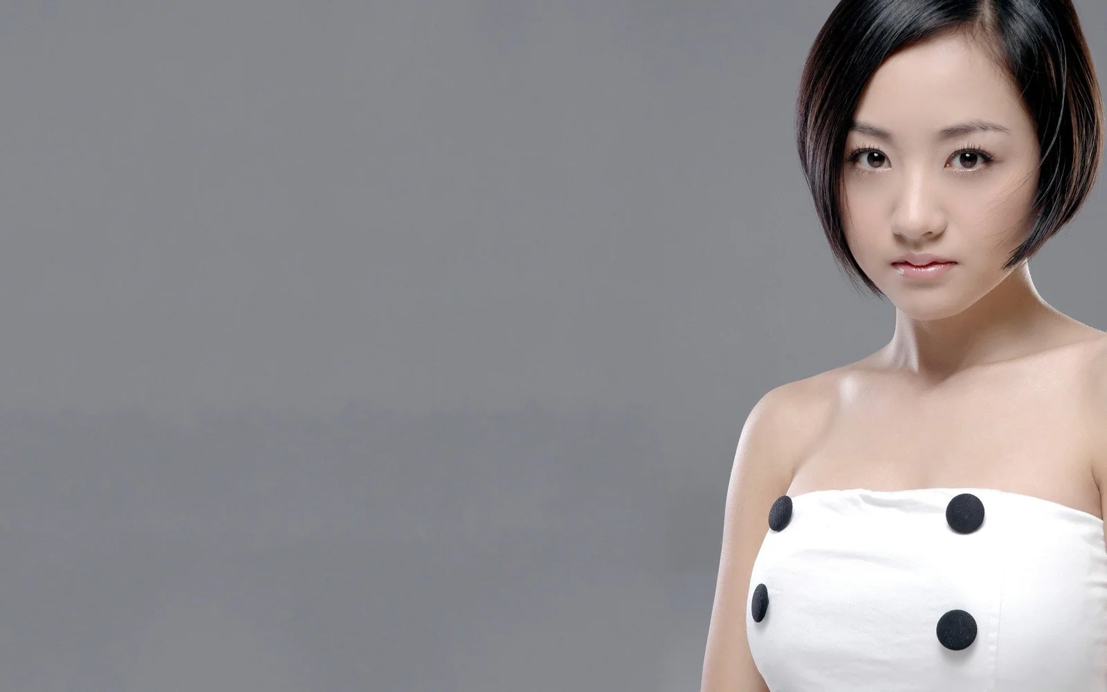 一位短发女性身穿白色无袖连衣裙站在灰色背景前。