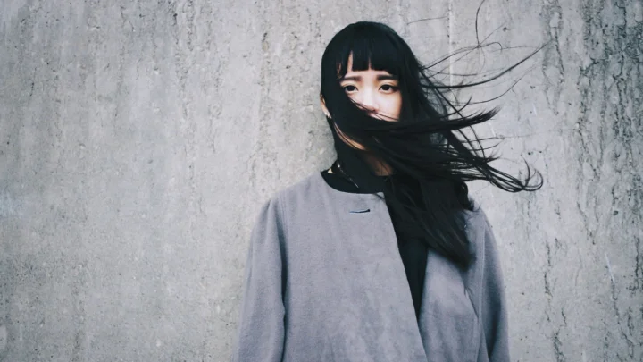 一名穿灰色外套的黑发女子站在灰色纹理墙前，长发被风吹向一侧。