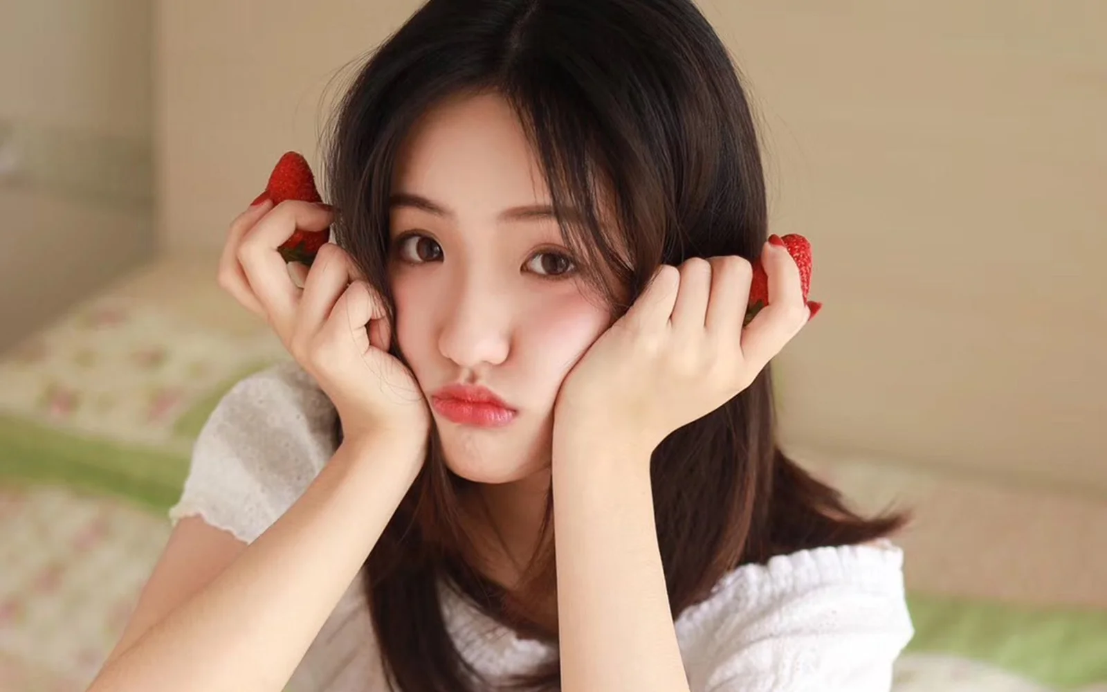긴 머리의 여성이 양손으로 볼을 괴고 얼굴 옆에 딸기를 든 채 카메라를 향해 장난스러운 표정을 짓고 있습니다.