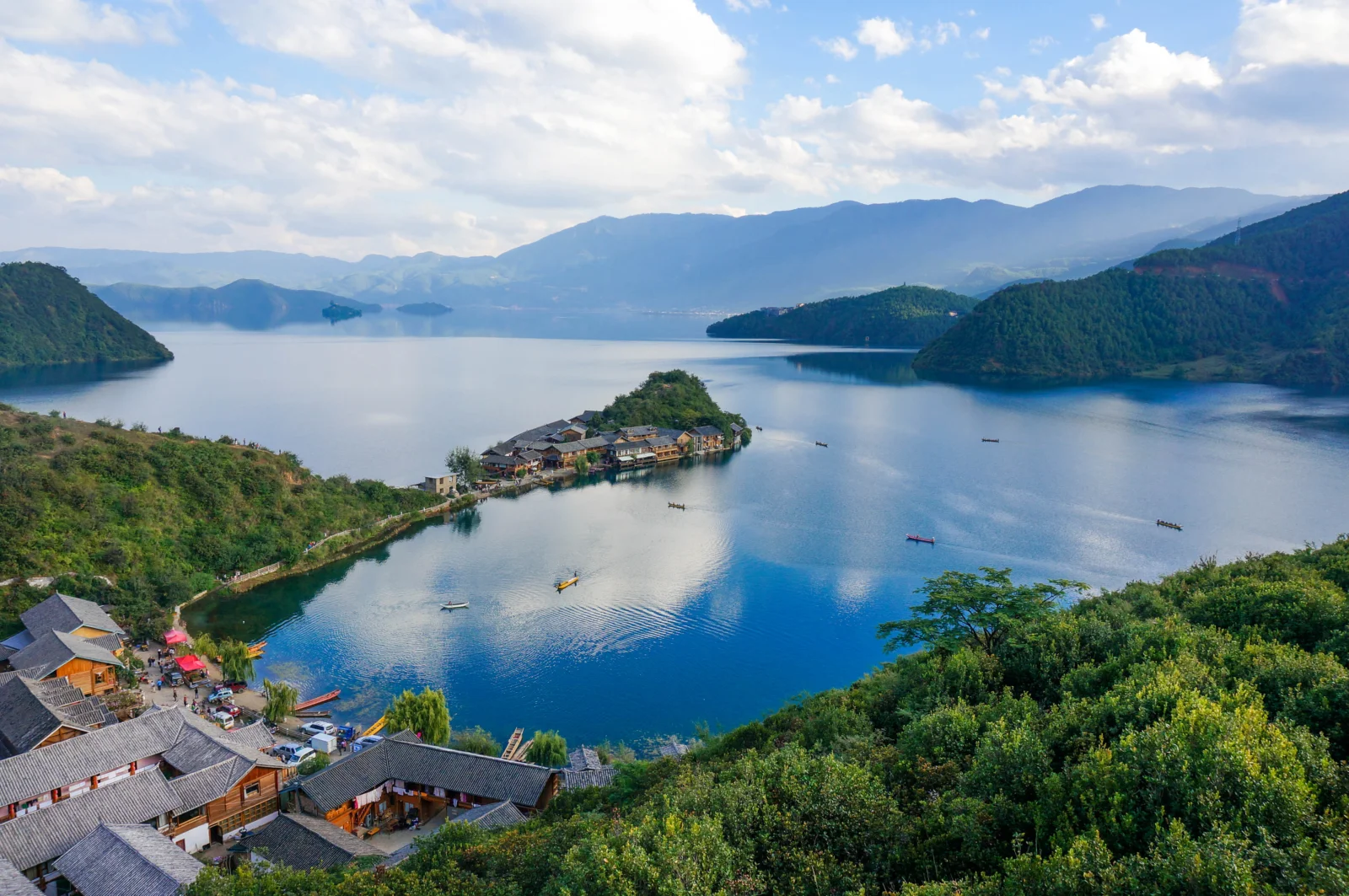 山々に囲まれた穏やかな湖が広がり、手前には森林や集落の屋根、水面の小舟が見える。