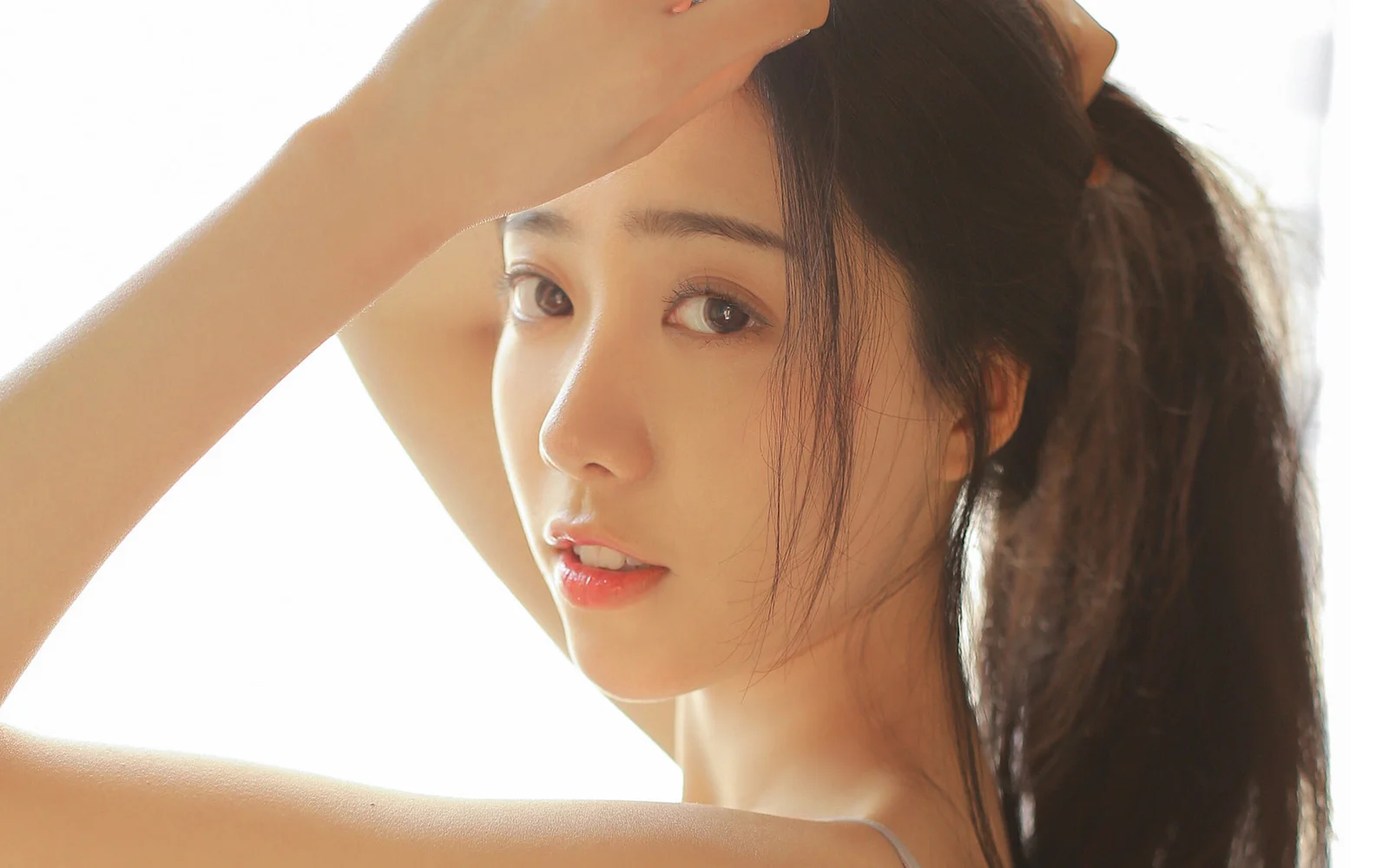 明るい背景の前で両腕を上げた、長い黒髪の成人女性のクローズアップポートレートです。