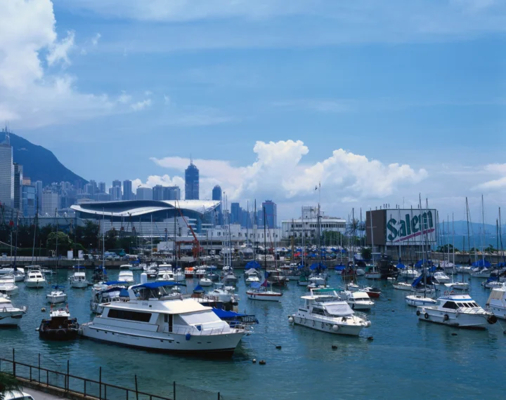 晴朗蓝天下的海港停泊着许多船只，远处是城市天际线和山脉。