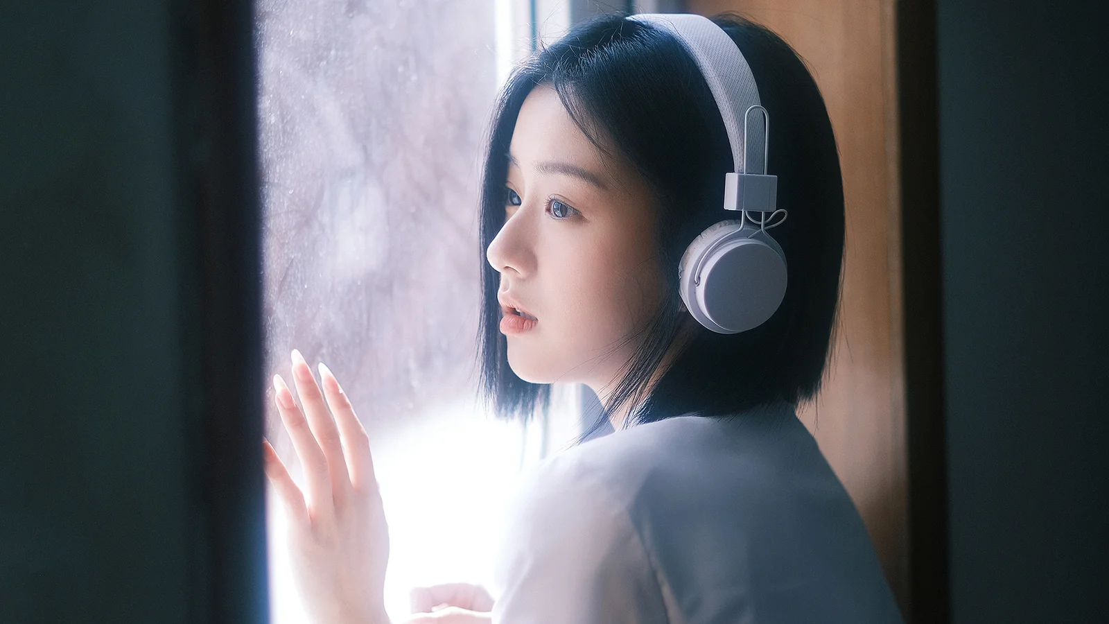 ヘッドホンを着けた女性が窓辺に立ち、窓の外から柔らかな光が差し込んでいる。