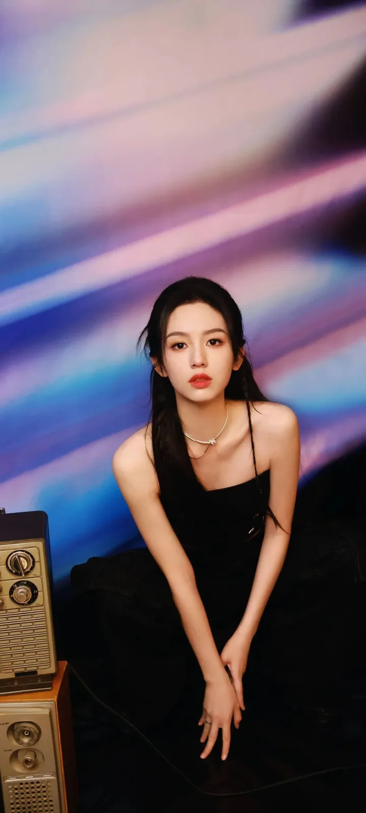 一名身穿黑色服裝的女性坐在彩色光影背景前。