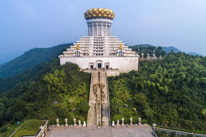 一座宏伟的古建筑坐落在郁郁葱葱的山林之间，远处可见层叠山峦。