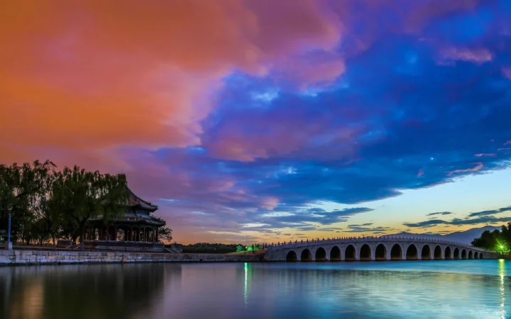 晚霞映照下的湖面旁，一座古典亭台与拱桥构成宁静的风景画面。