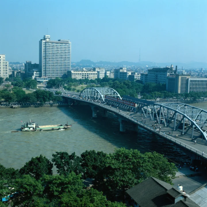 一座钢架桥横跨宽阔河流，背景是城市建筑与远处山丘。