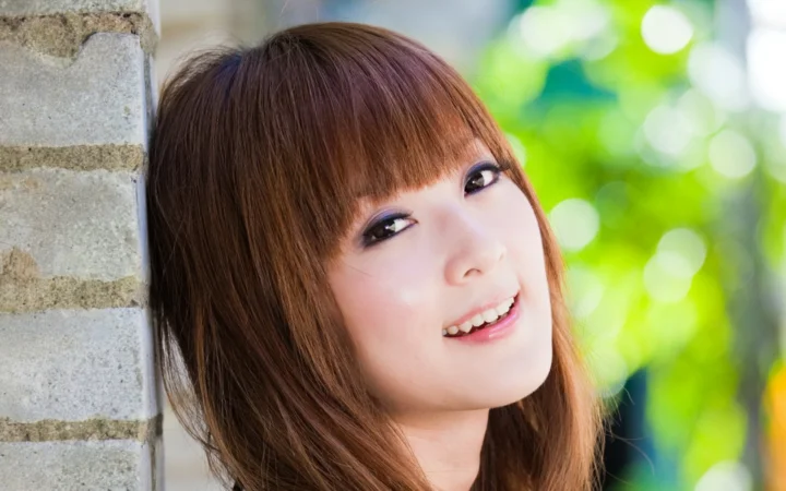茶色の長い髪の女性が、明るい緑の屋外背景を前にレンガの壁のそばで微笑んでいる。