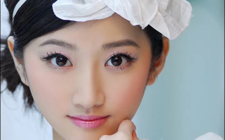 白いリボンを身につけた黒髪の女性が、淡い青色の背景の前で手を顎に添えたクローズアップポートレート。