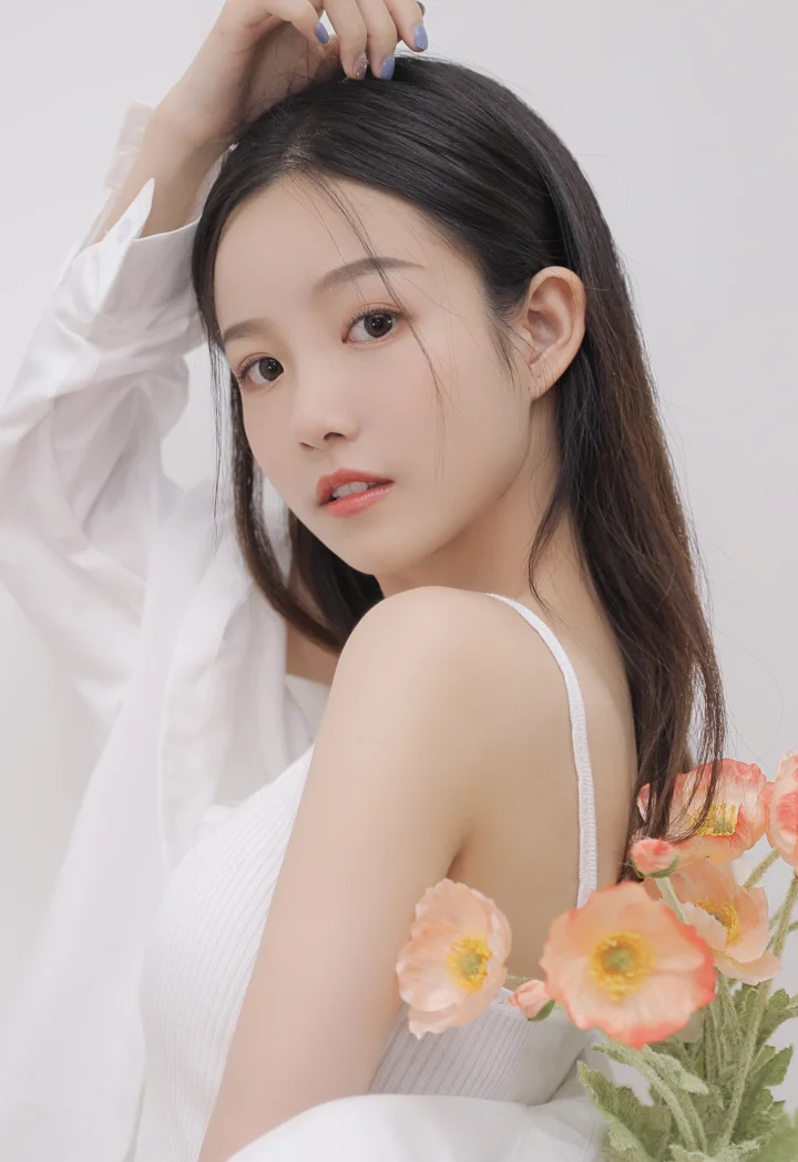 흰색 상의를 입은 성인 여성이 밝은 배경 앞에서 뒤돌아보며 바라보고 있고, 오른쪽 아래에는 분홍색 꽃이 장식되어 있습니다.