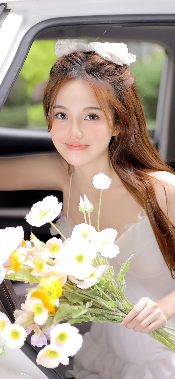 一名长发女子坐在车内，手捧一束白色和黄色花朵。